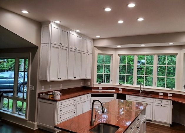 A bright kitchen featuring white cabinets, a red granite island with a sink, and large windows overlooking green trees.