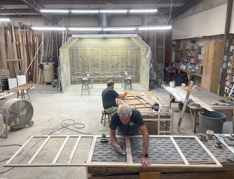 Two people work on wood frames in a workshop with a large spray booth in the background.
