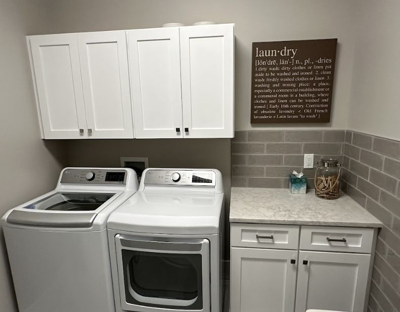White laundry room with washer, dryer, beige cabinets, gray subway tile backsplash, and a wall sign.