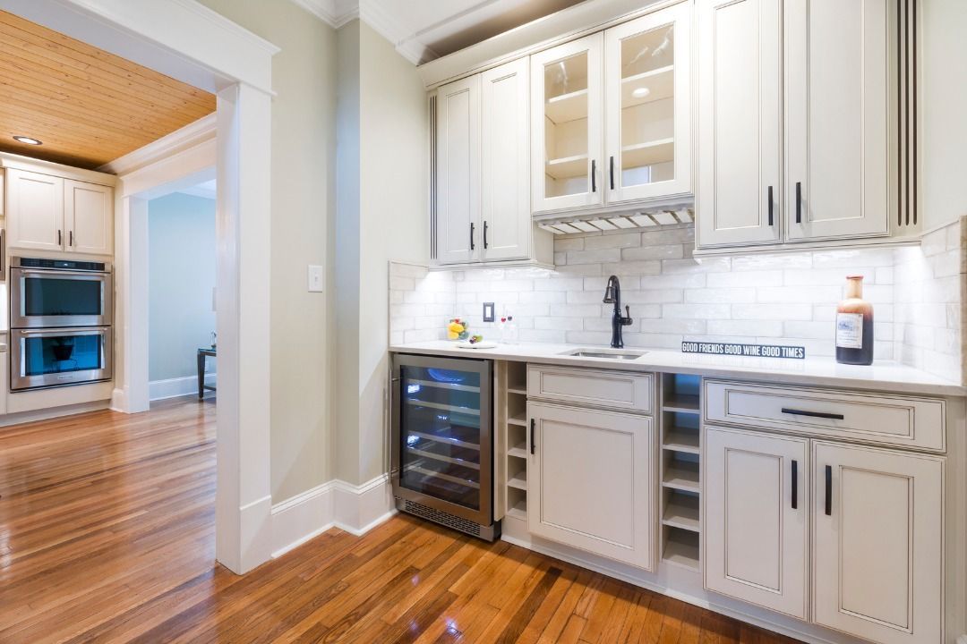 A wet bar with white cabinets, a wine fridge, glass shelving, and a small sink against a white subway tile backsplash.