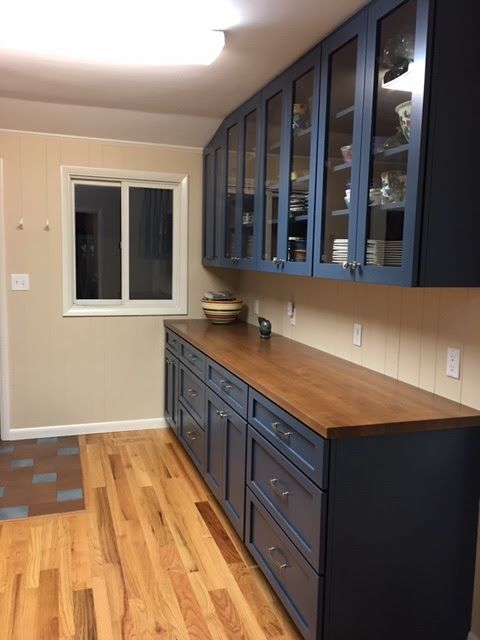 Dark blue cabinets with glass uppers and a wooden countertop line a kitchen wall next to a window and hardwood floors.