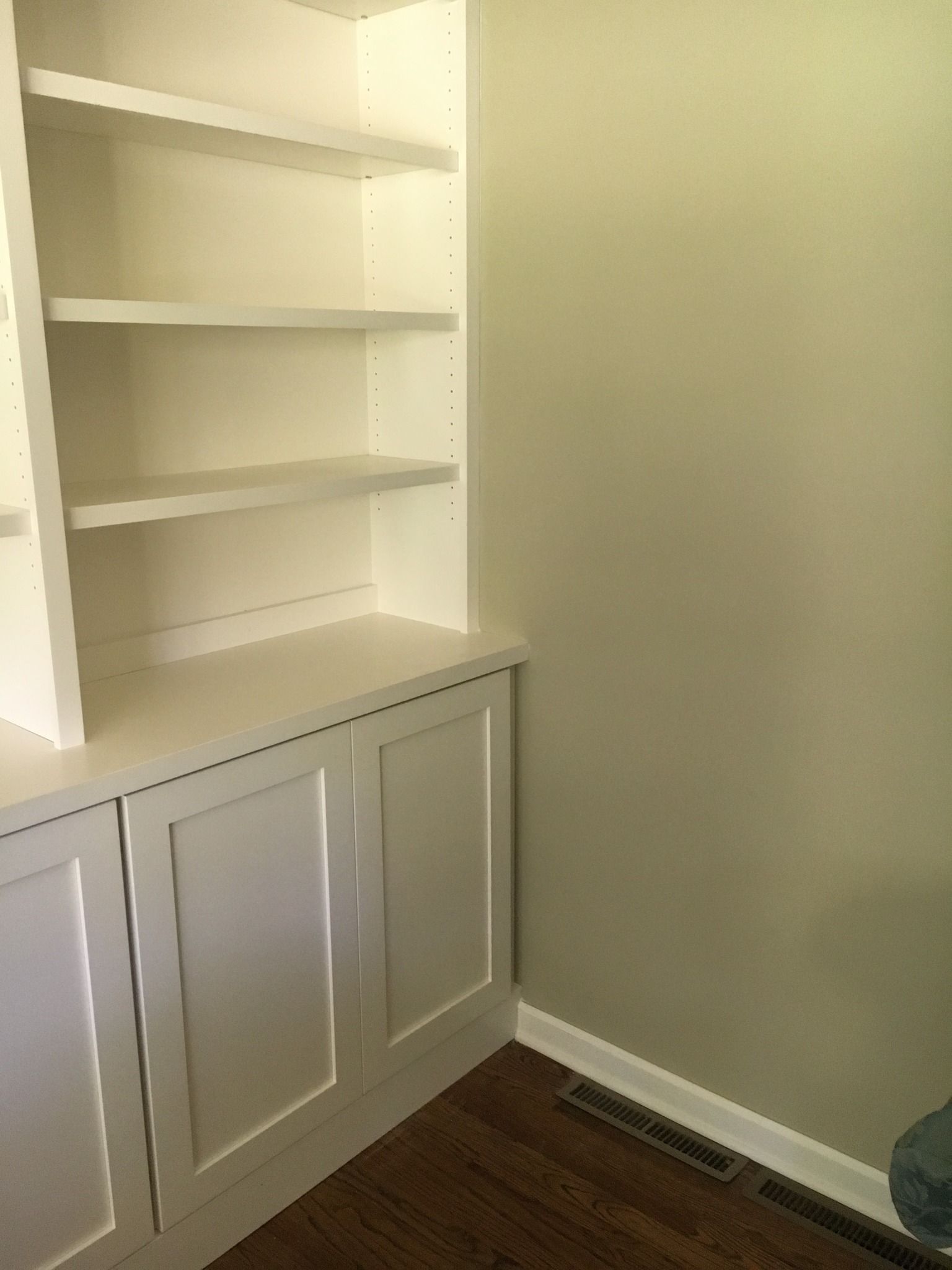 A white built-in bookshelf and base cabinet unit set against a pale green wall with dark wood flooring.