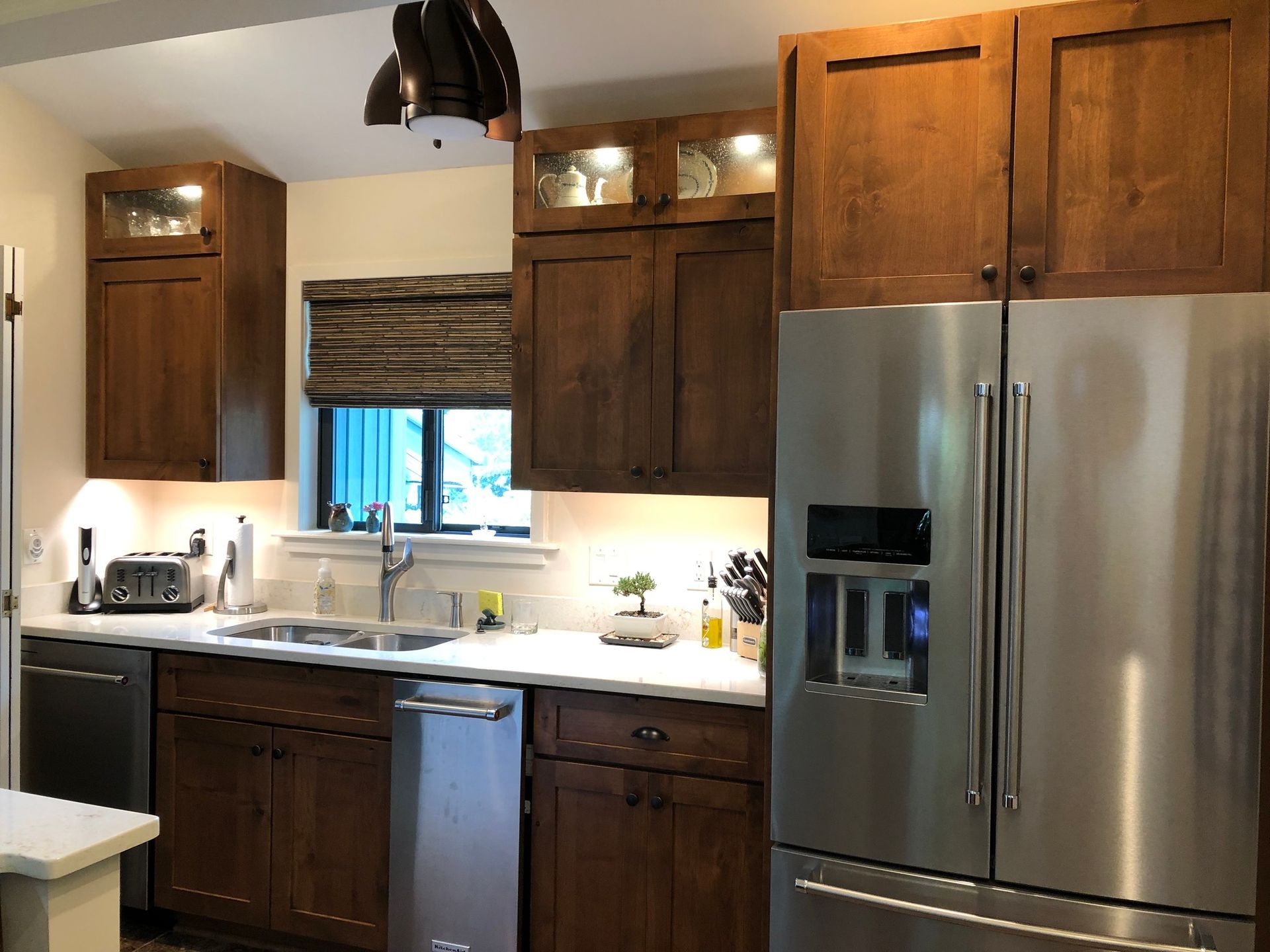 A kitchen with dark wood cabinets, a stainless steel refrigerator and dishwasher, and a window with a woven shade.