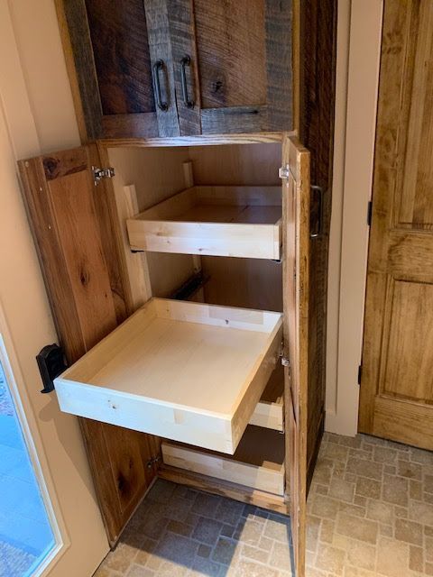 A rustic wooden pantry cabinet with open doors showing light-colored pull-out shelving drawers.