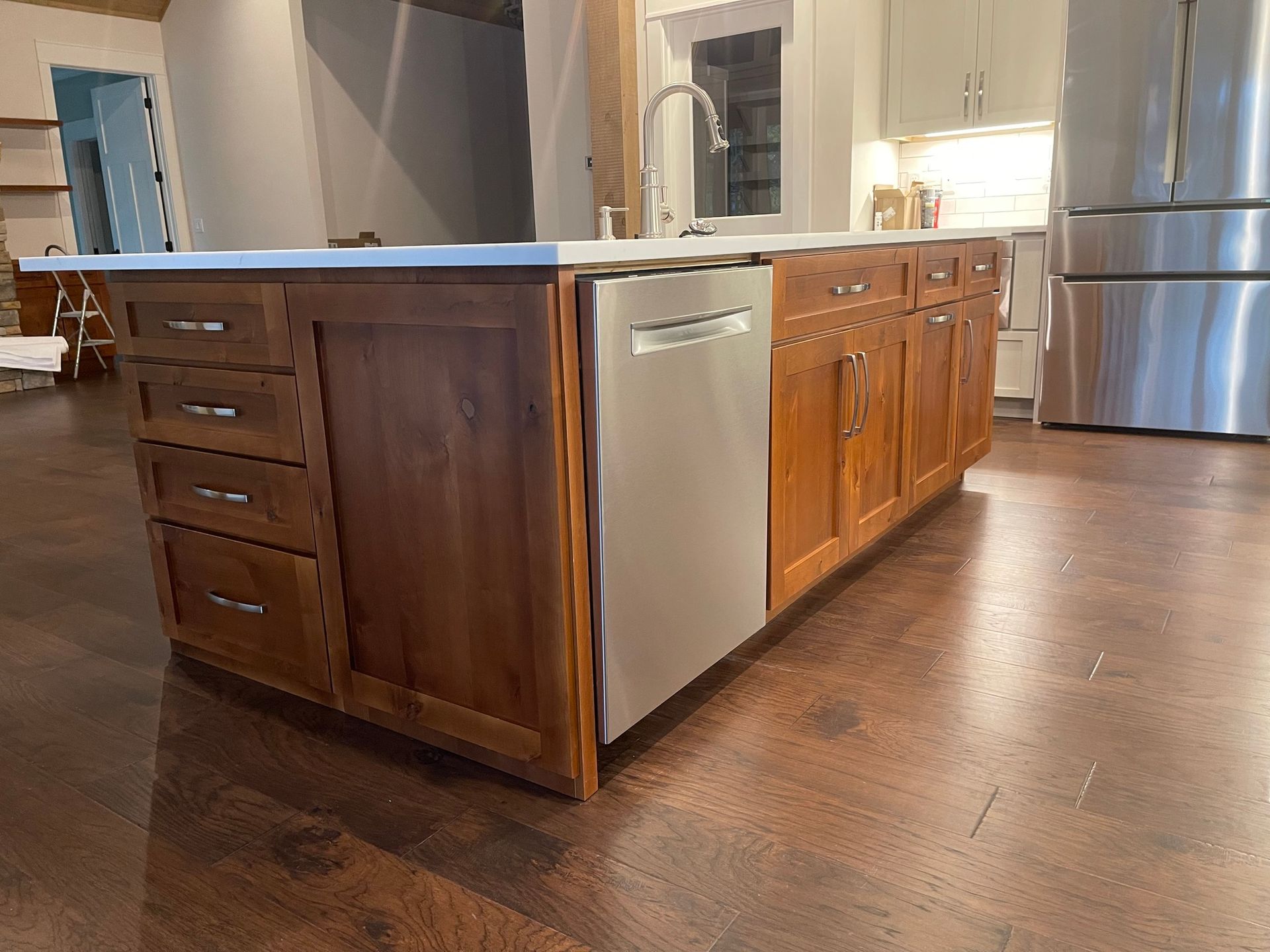 A kitchen island with warm-toned wooden cabinets, a stainless steel dishwasher, and a white countertop on a wood floor.
