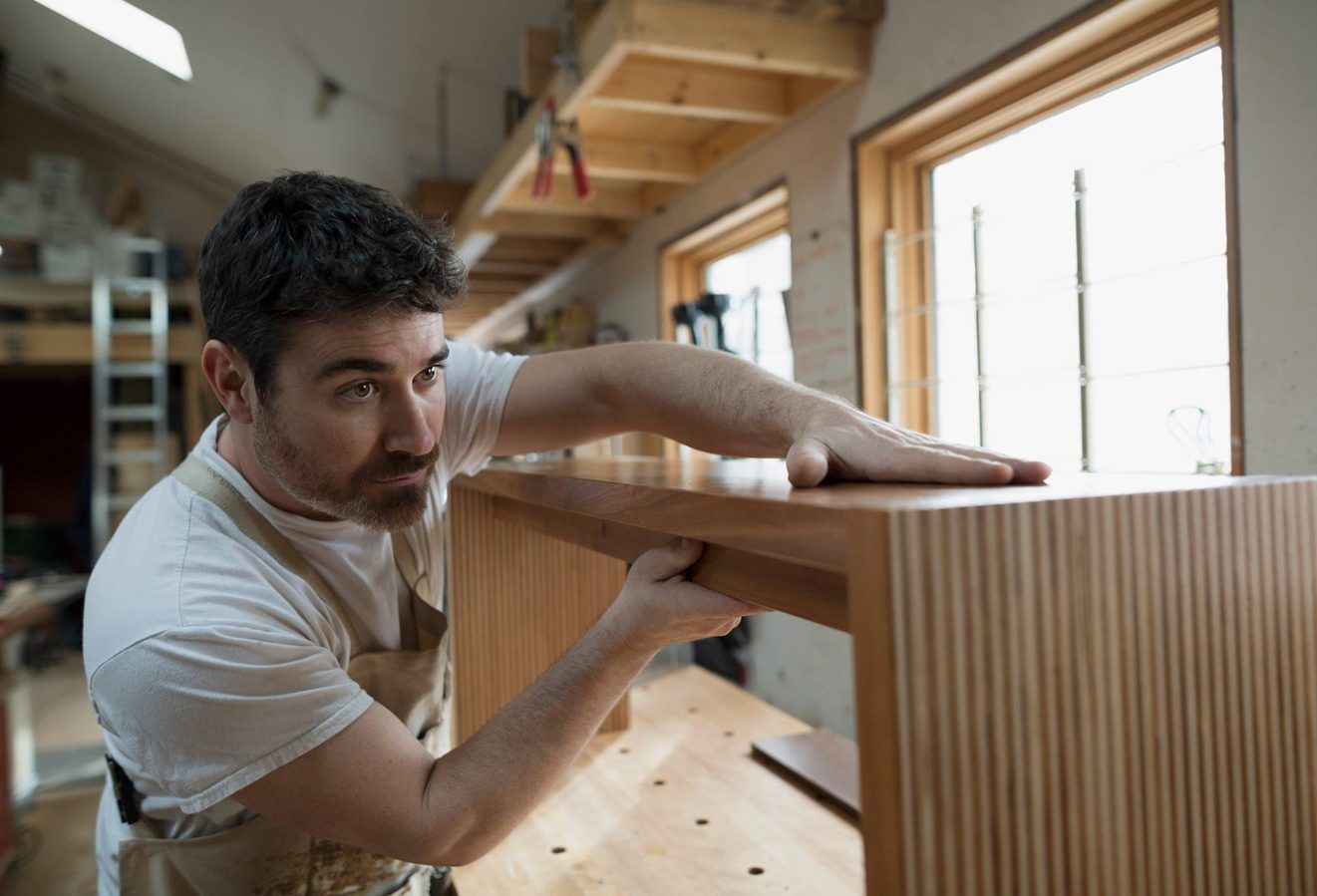 A woodworker in a workshop inspects the smooth surface of a custom wooden furniture piece.