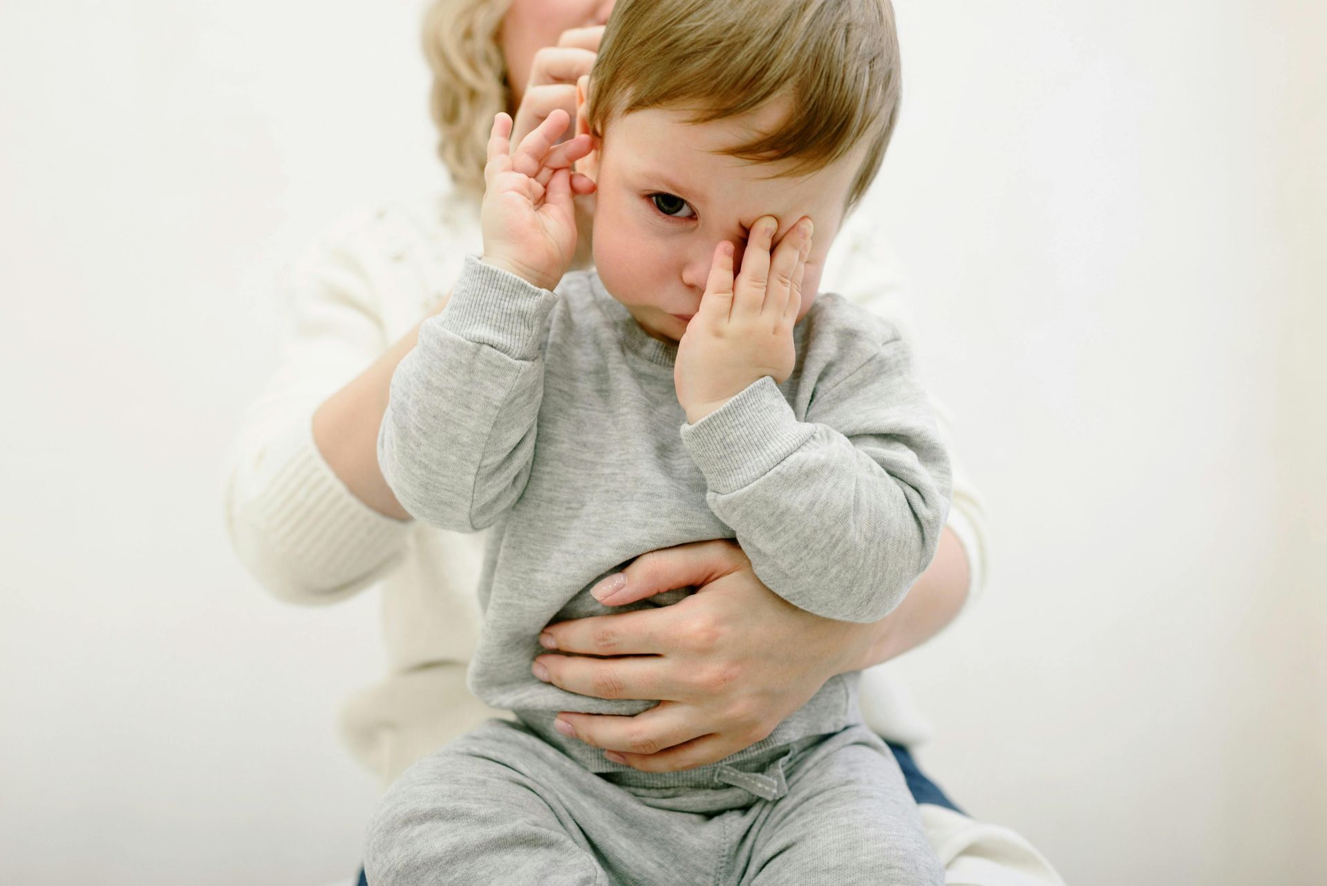 A sick boy rubs his face while his mom holds him