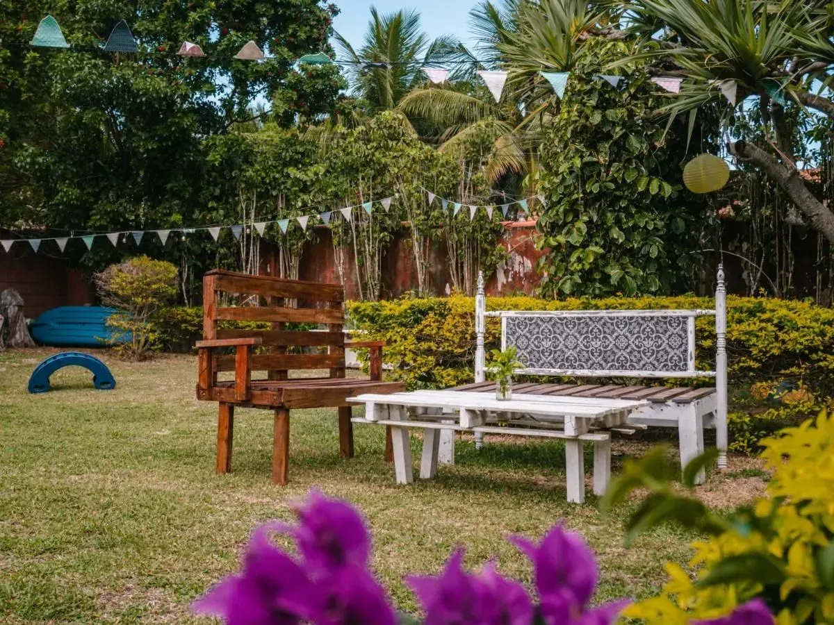 Jardín con bancos de madera y blancos sobre un césped, con plantas tropicales y flores moradas en primer plano.