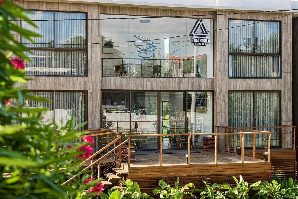 Edificio moderno de hormigón con grandes ventanales y una terraza de madera, rodeado de vegetación.