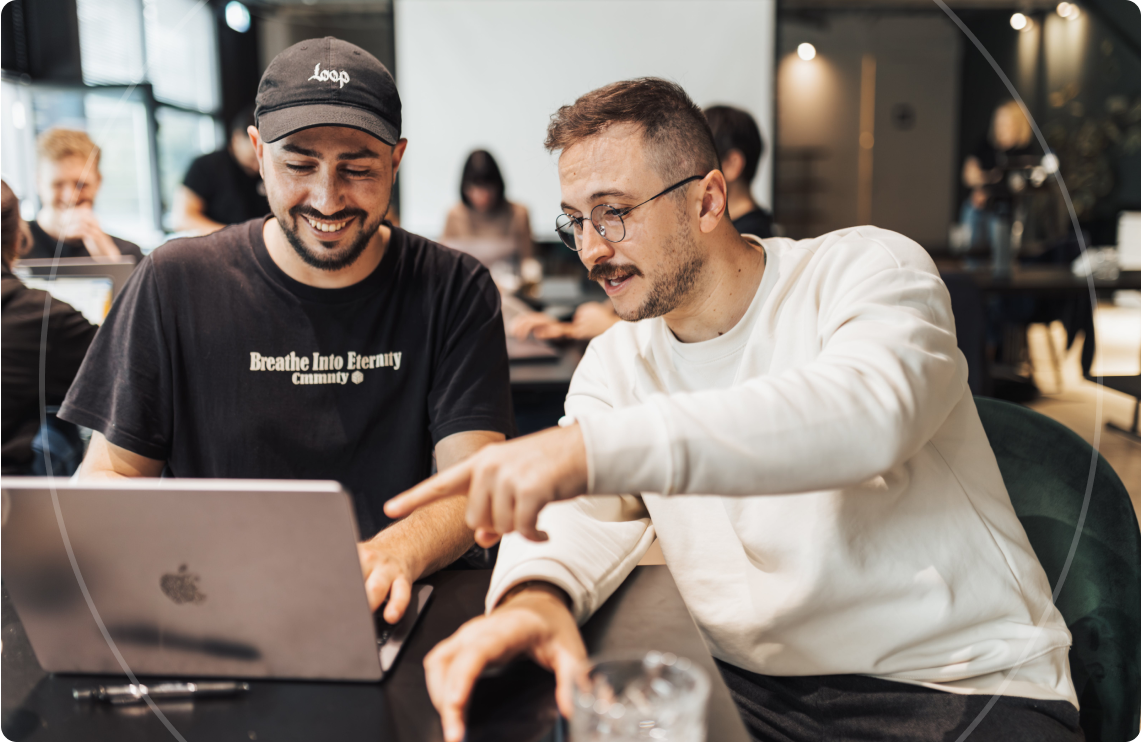 Dos hombres sonriendo y señalando una computadora portátil en un entorno de oficina informal.
