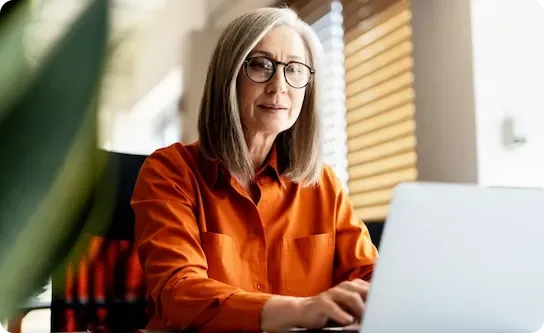Pessoa vestindo camisa laranja digitando em um laptop em um escritório bem iluminado com persianas.