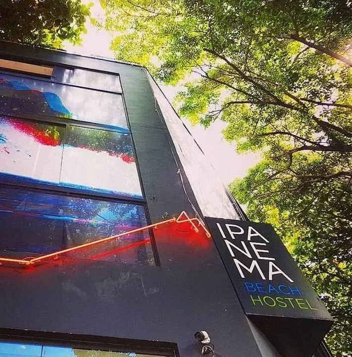 Edificio moderno de cristal con un letrero de PANE MAE HOSTEL, visto desde abajo a través de árboles frondosos.