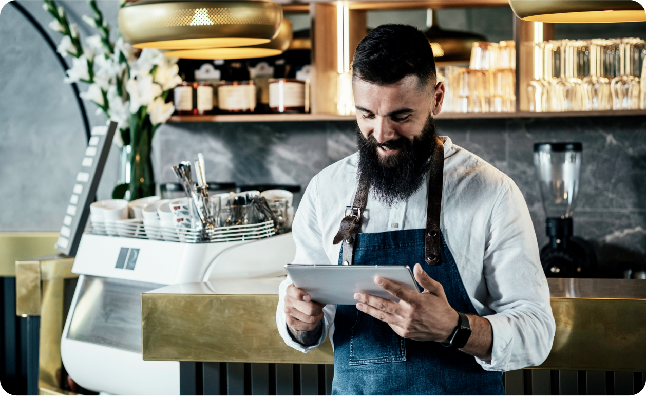 Barista con delantal usando una tableta detrás de la barra de una cafetería con máquina de espresso y vasos al fondo.