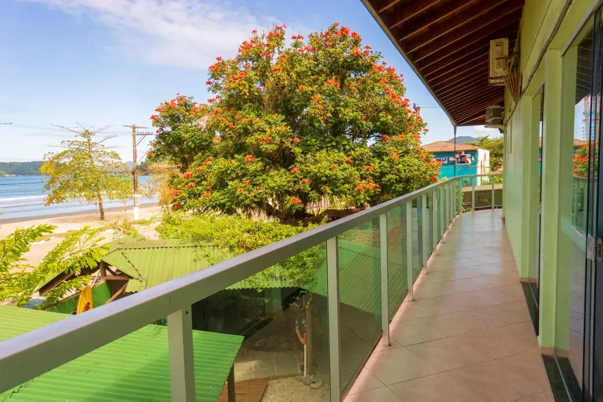 Balcón con barandilla de cristal con vistas a una playa tropical y un árbol en flor junto a un edificio con techo verde.