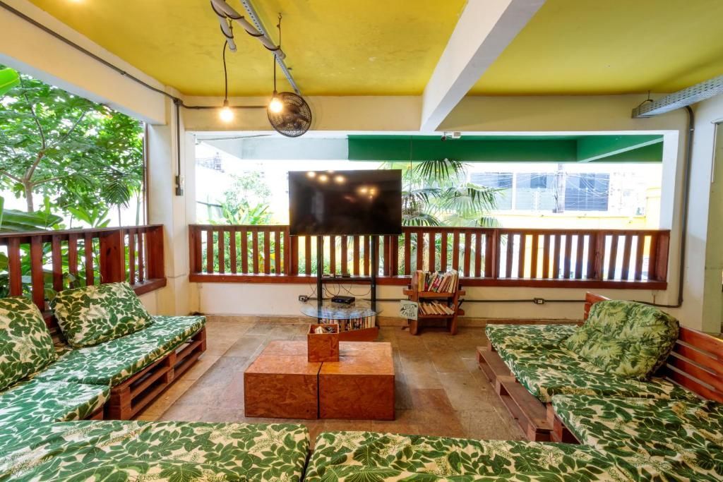 Terraza cubierta con sofás de estampado verde, televisor y barandilla de madera con vistas a la vegetación.