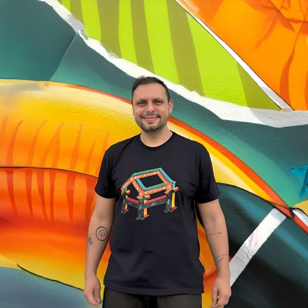 Un hombre, de pie frente a un colorido mural abstracto, viste una camiseta negra y sonríe.