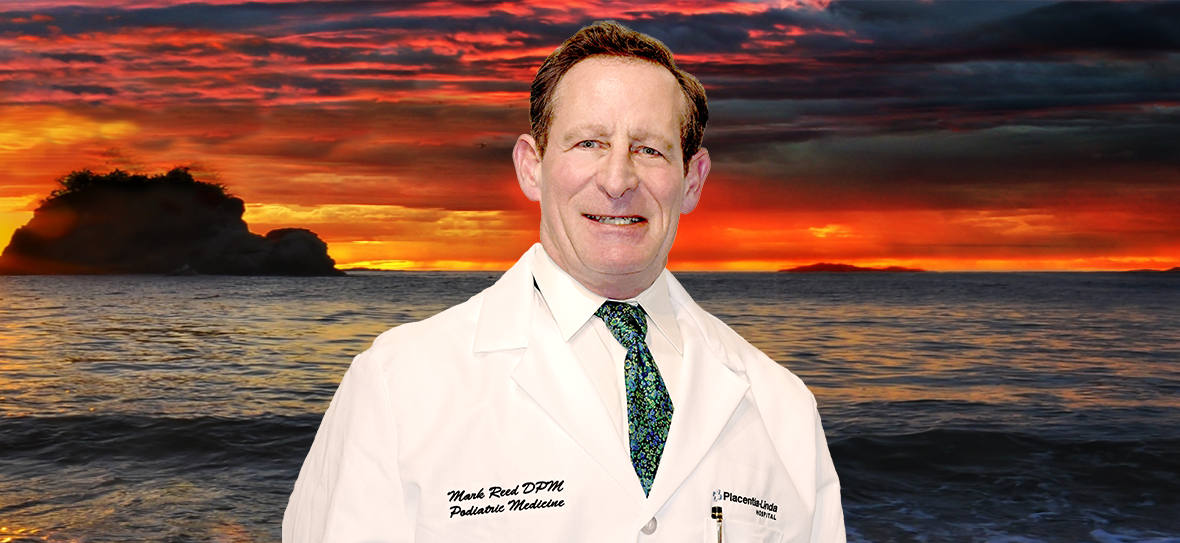 A man in a white coat and tie is standing in front of a sunset over the ocean.