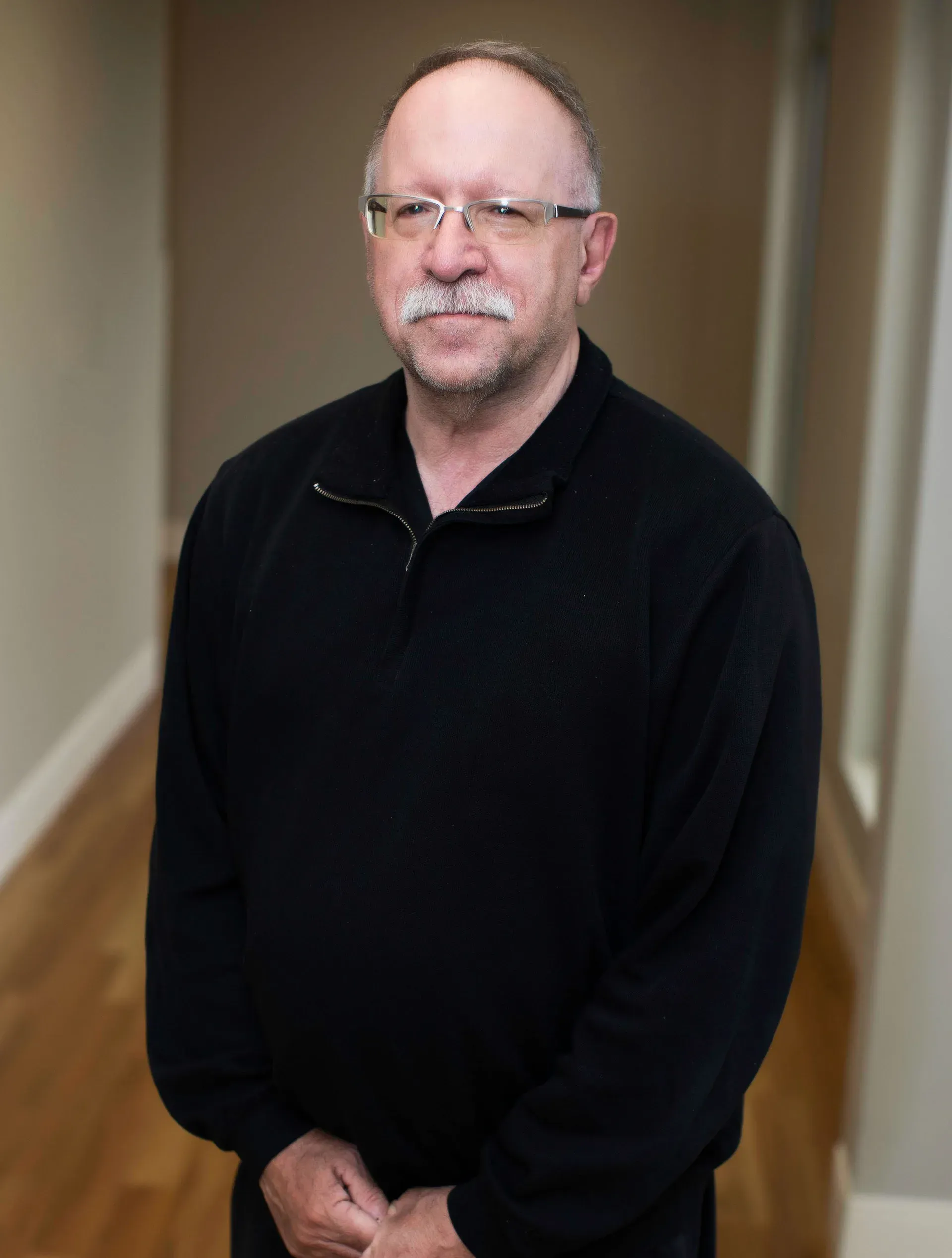 Man with glasses and a mustache in a black sweater, standing in a hallway with wood flooring.