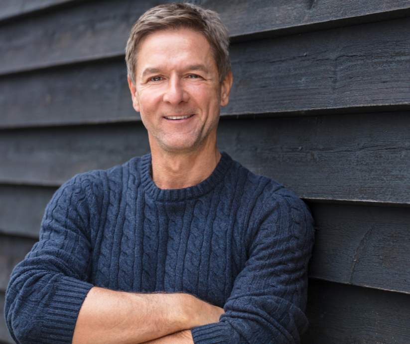 Man in blue sweater smiles, arms crossed, leaning against a dark wood wall.