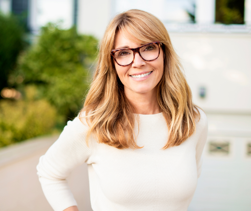 Woman with shoulder-length blonde hair and glasses smiles, posing outside a building in front of greenery.