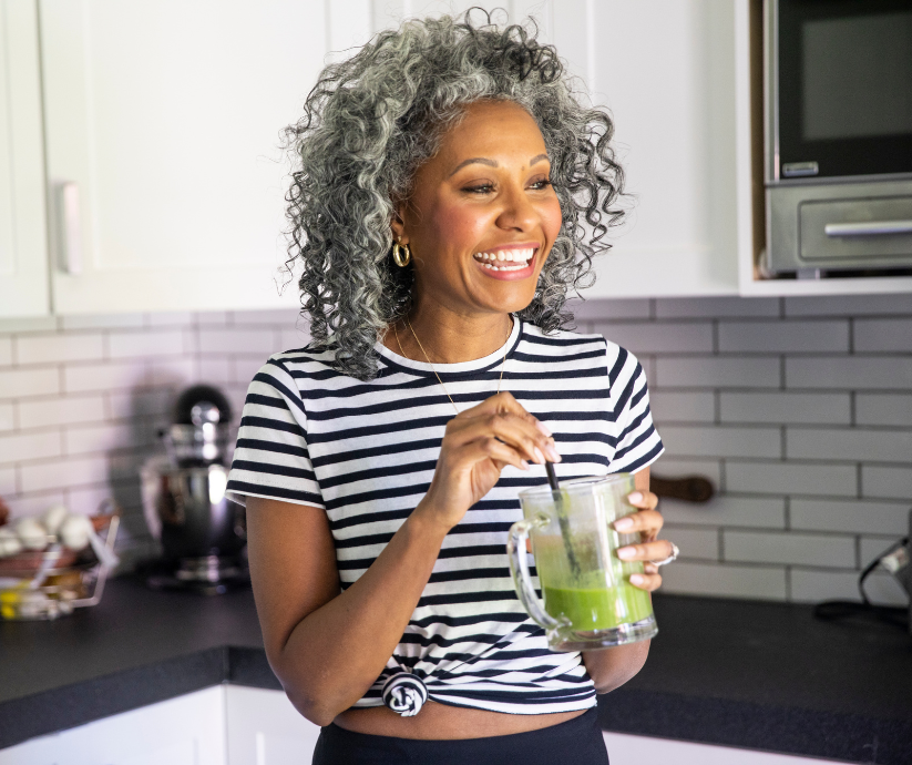 Woman with curly gray hair smiles, sipping a green drink in a kitchen.