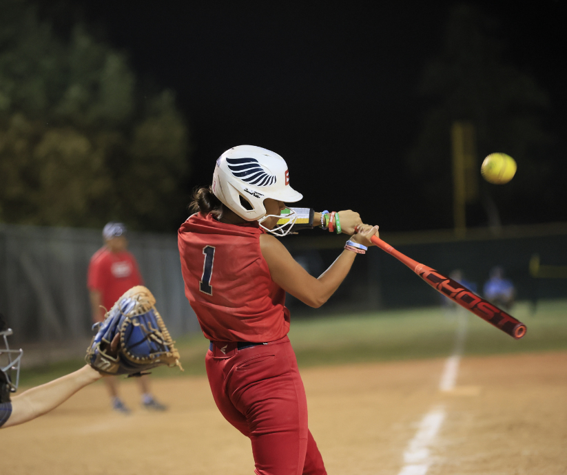 Softball player in red uniform swings bat, hitting the yellow ball at night.