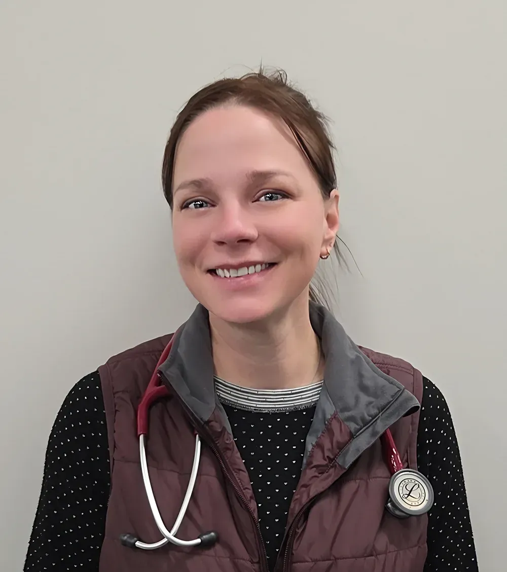 Woman with stethoscope, smiling, wearing a vest and sweater. Against a neutral background.