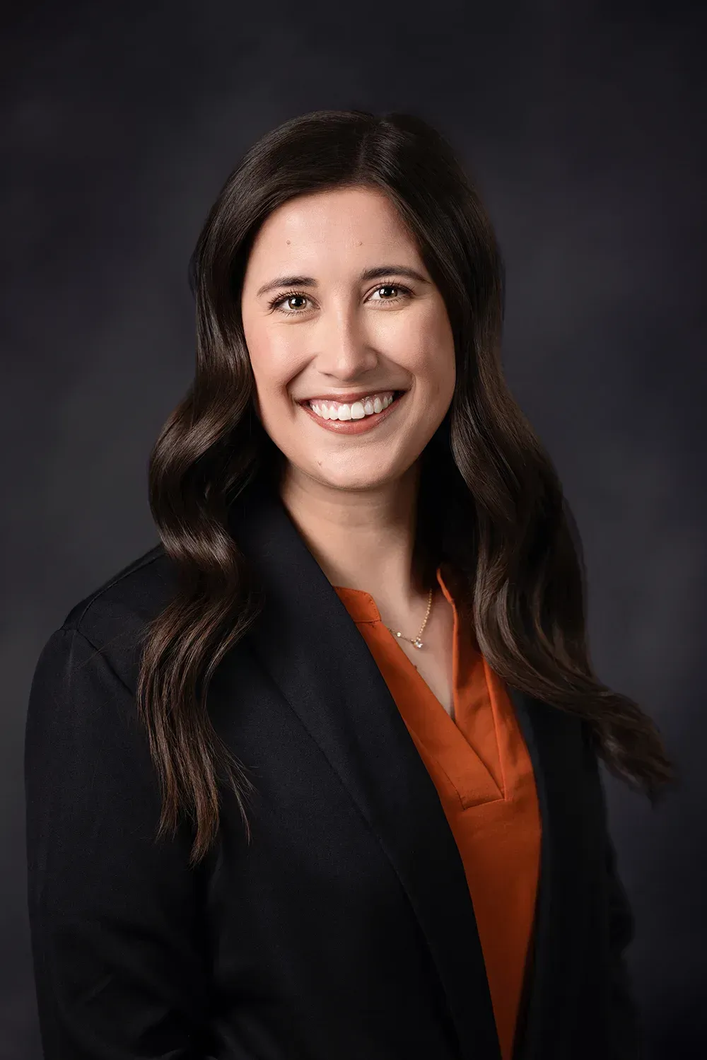 Woman with long brown hair, smiling, wearing a dark blazer and orange shirt, against a dark gray backdrop.