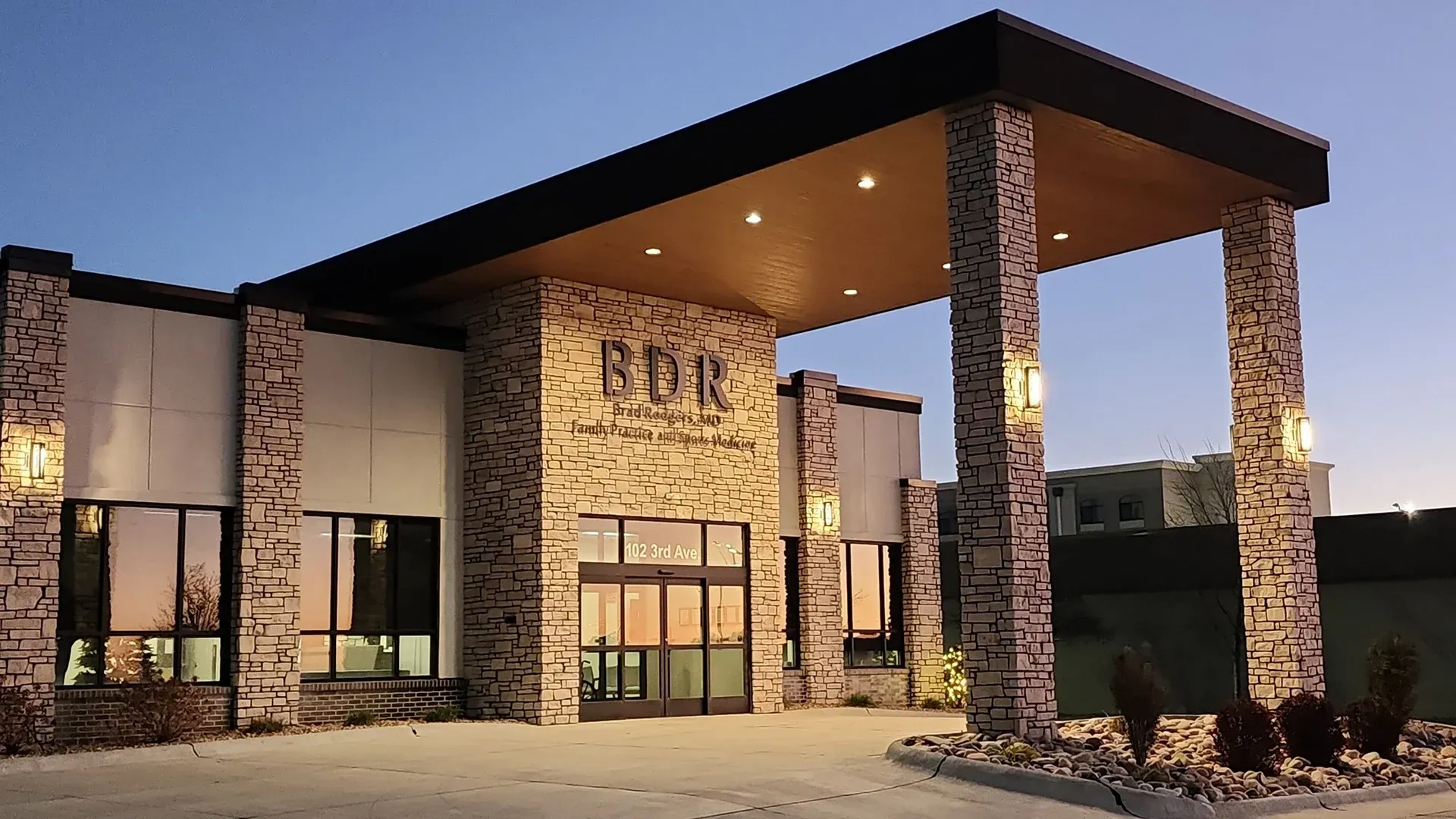 Exterior of a modern building with stone columns, glass windows, and a covered entrance at dusk.