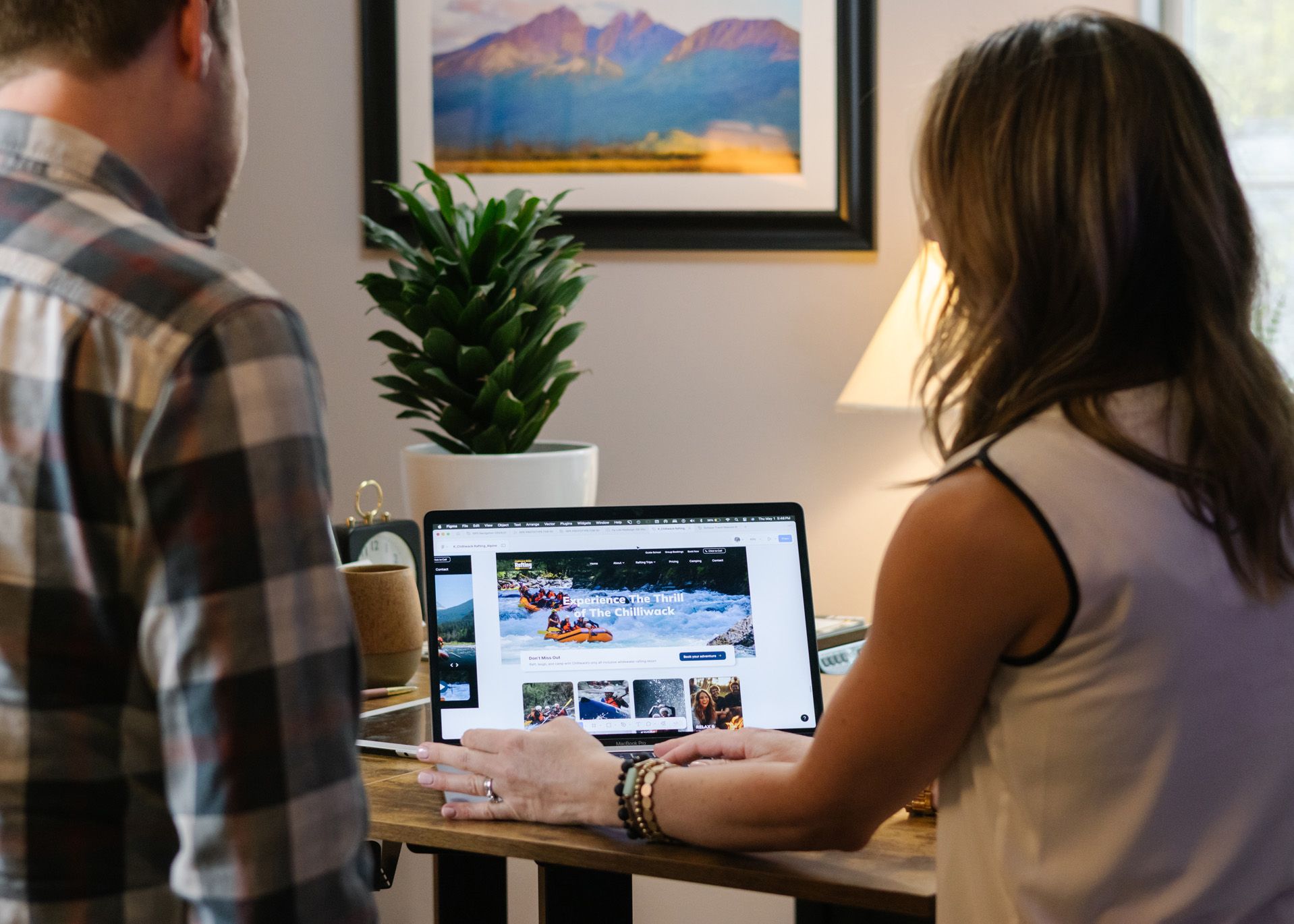 Couple looking at a laptop displaying photos of people rafting, indoors, near a plant and framed artwork.