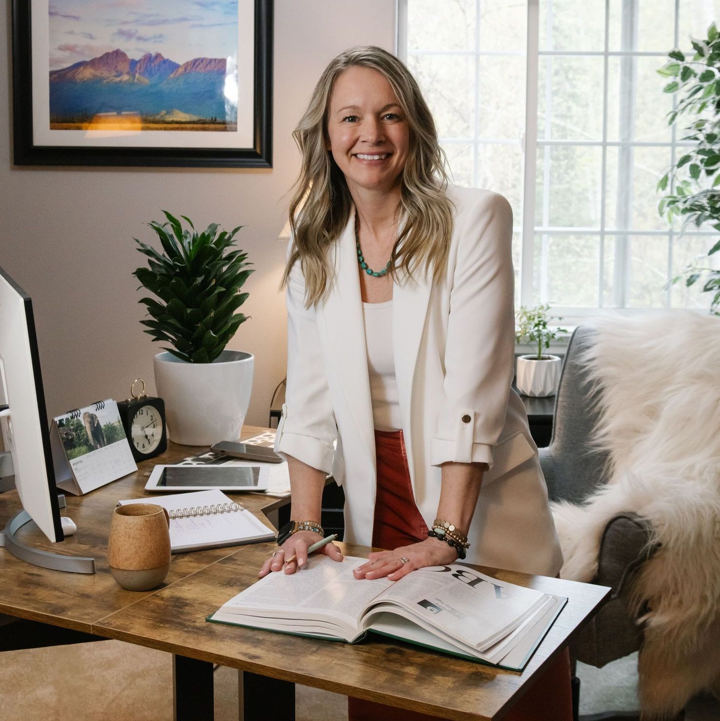 Kristin Kniss in white blazer stands at desk with open book, smiling.