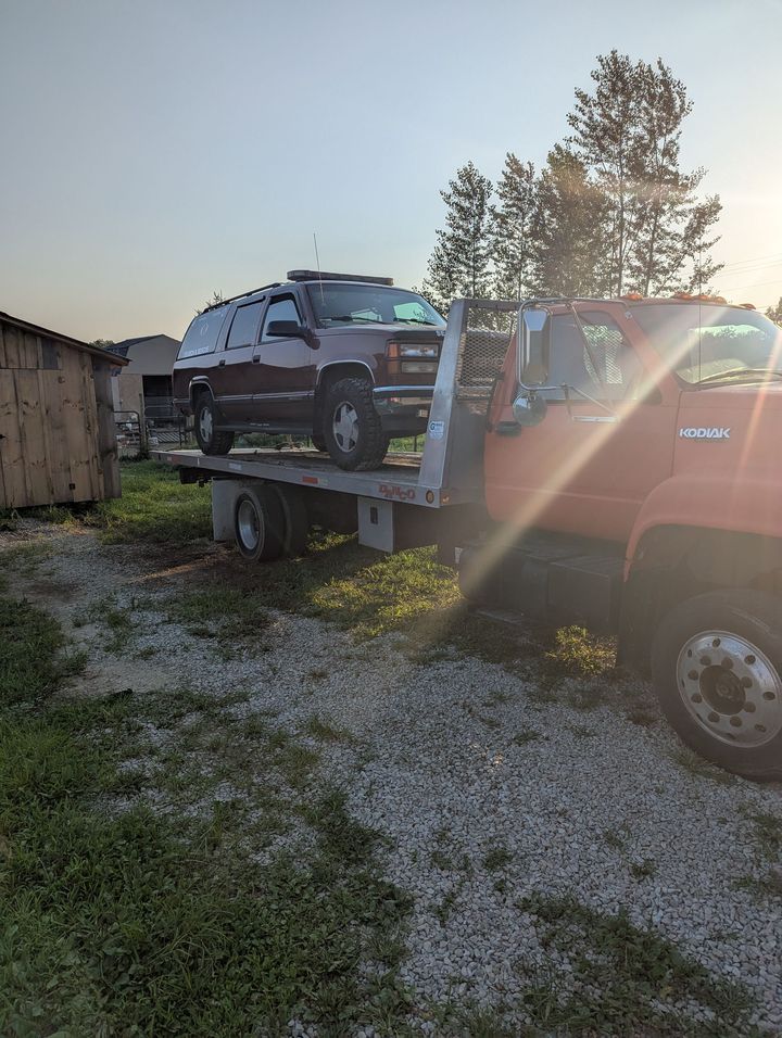 SUV on a flatbed tow truck in a gravel lot at sunset, with lens flare and trees in the background