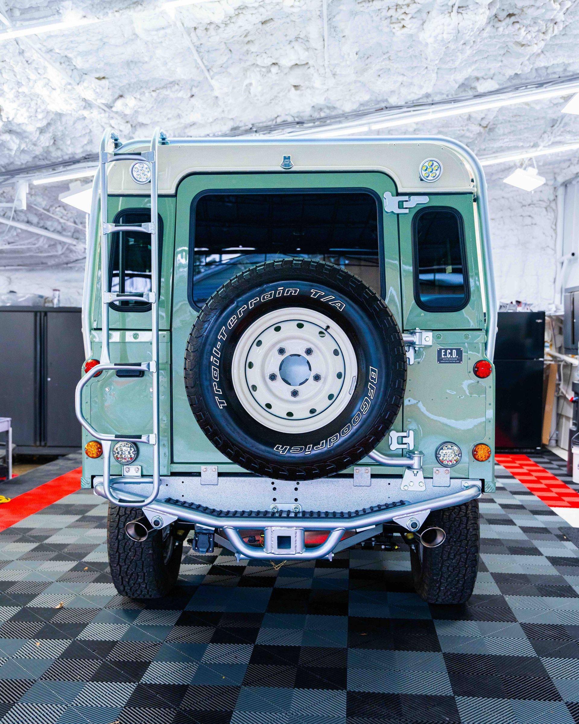 Rear view of a light green off-road vehicle with a spare tire, parked indoors on checkered flooring.