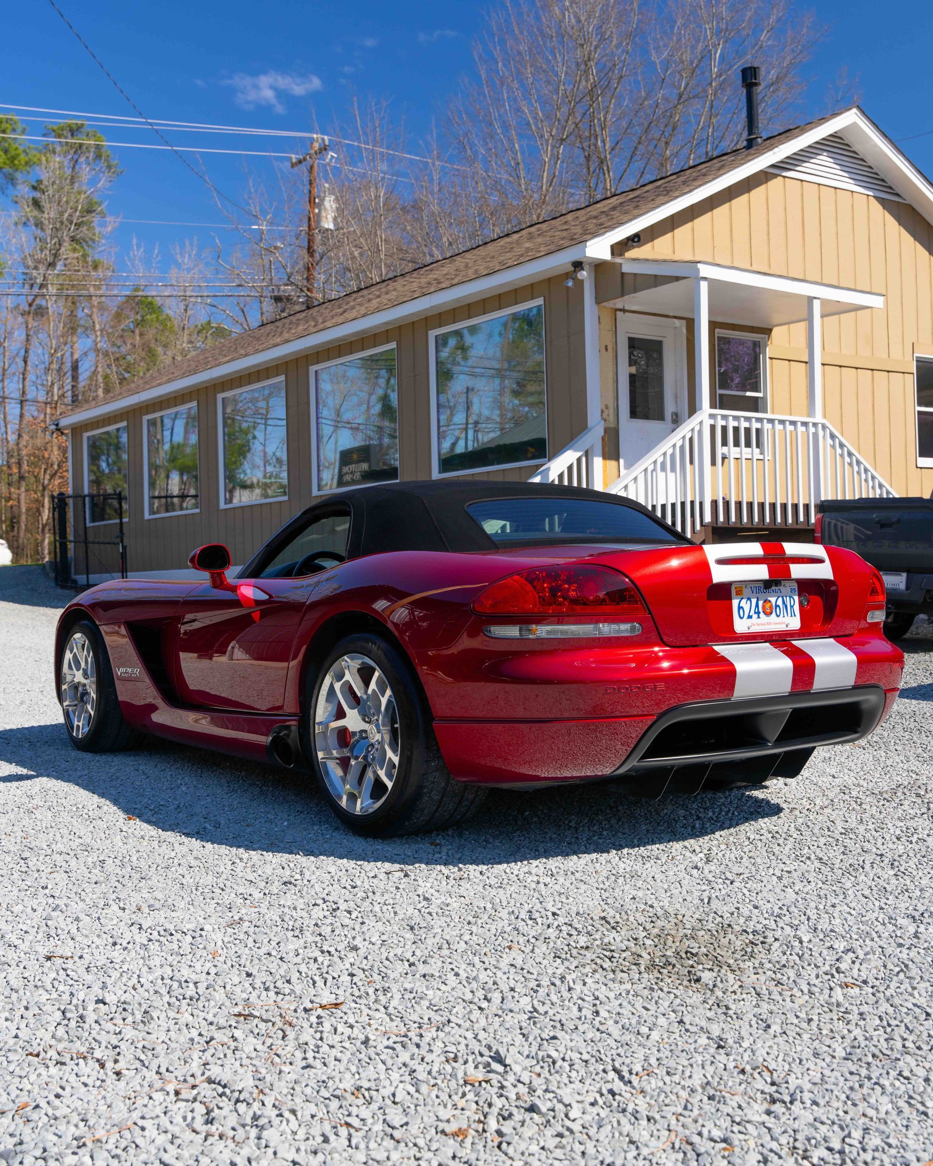 Red sports car with white racing stripes parked on gravel beside a tan house under a blue sky