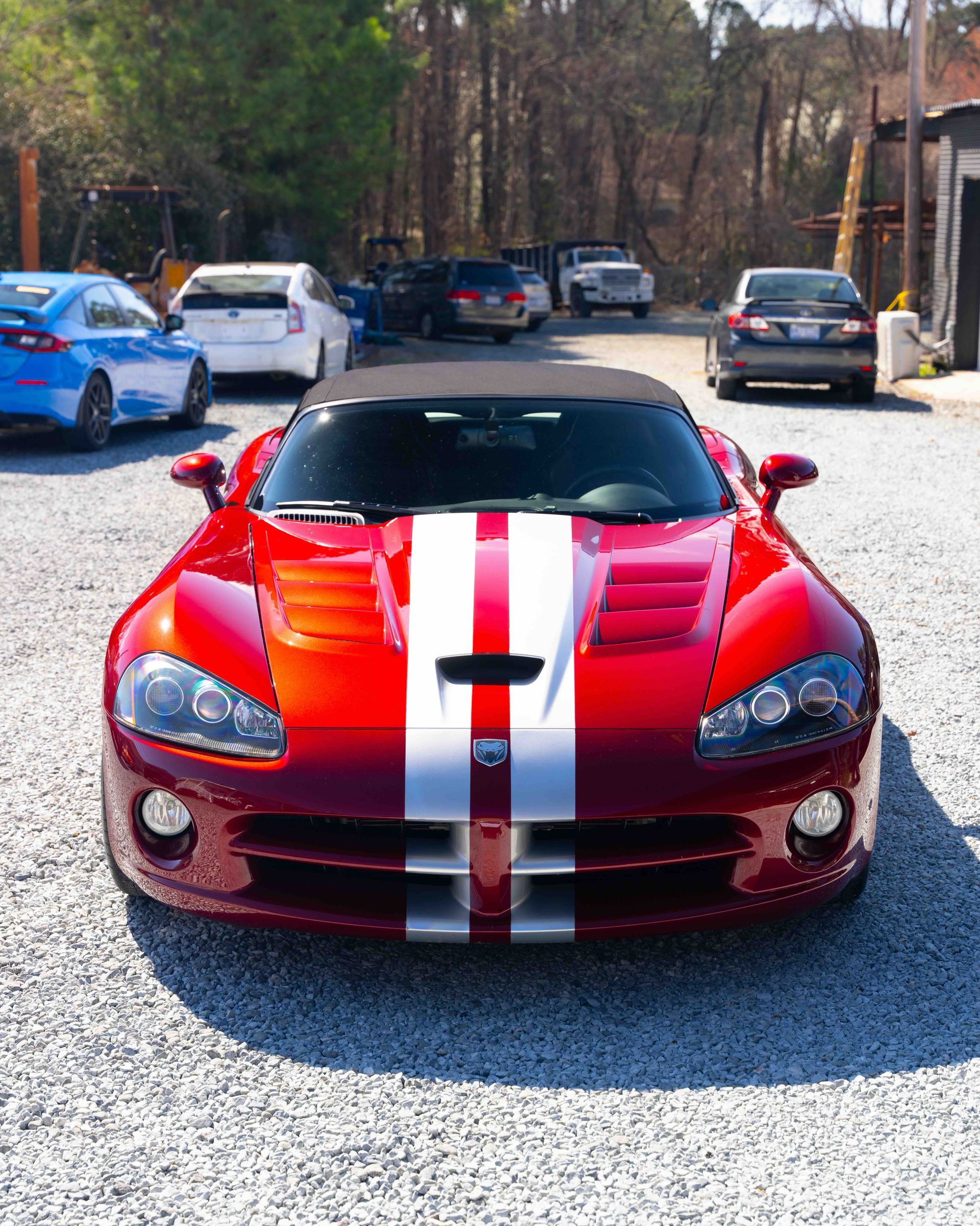 Red sports car with white racing stripes parked on a gravel lot, front view.