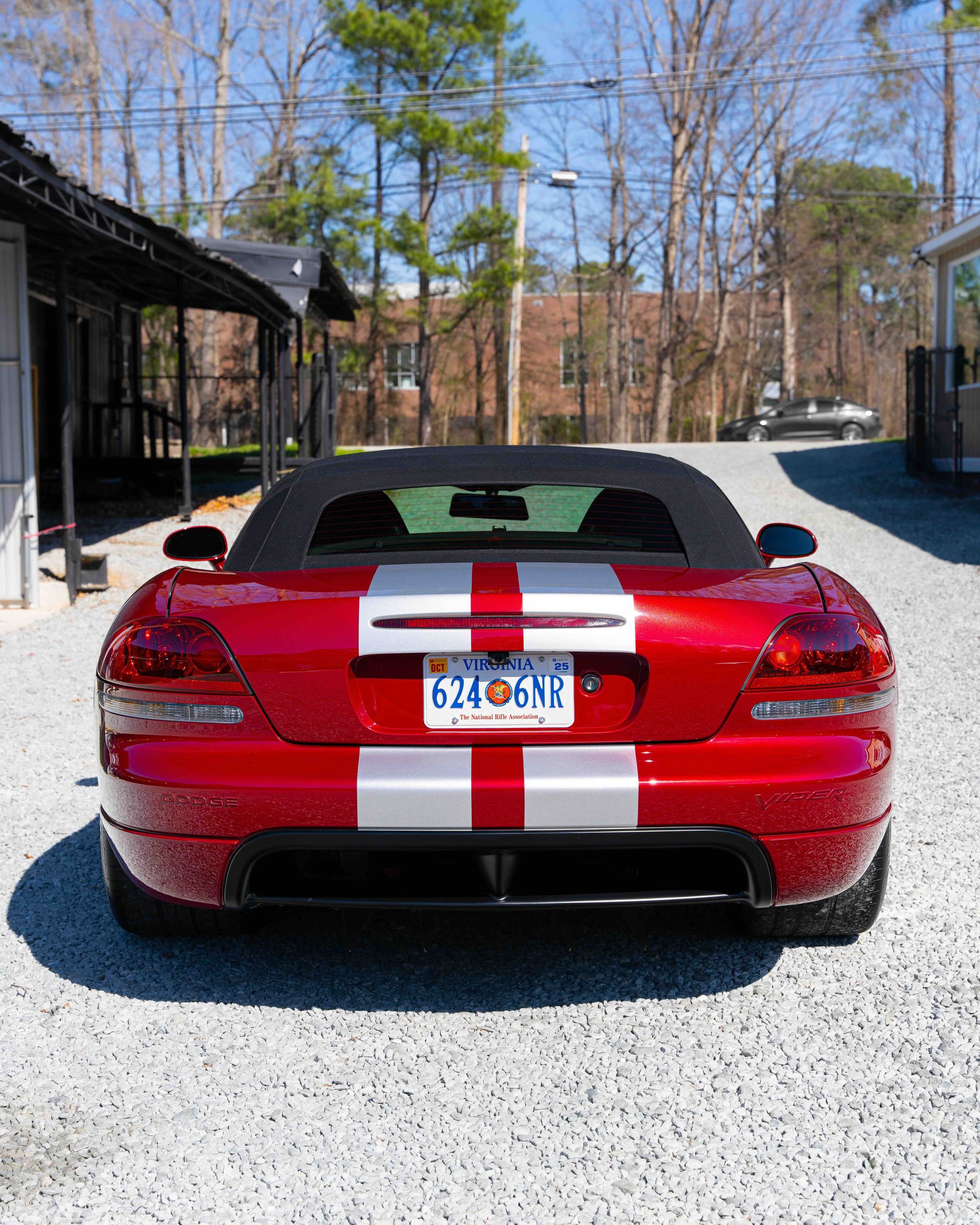 Red sports car with white racing stripes parked on gravel in a driveway between buildings.