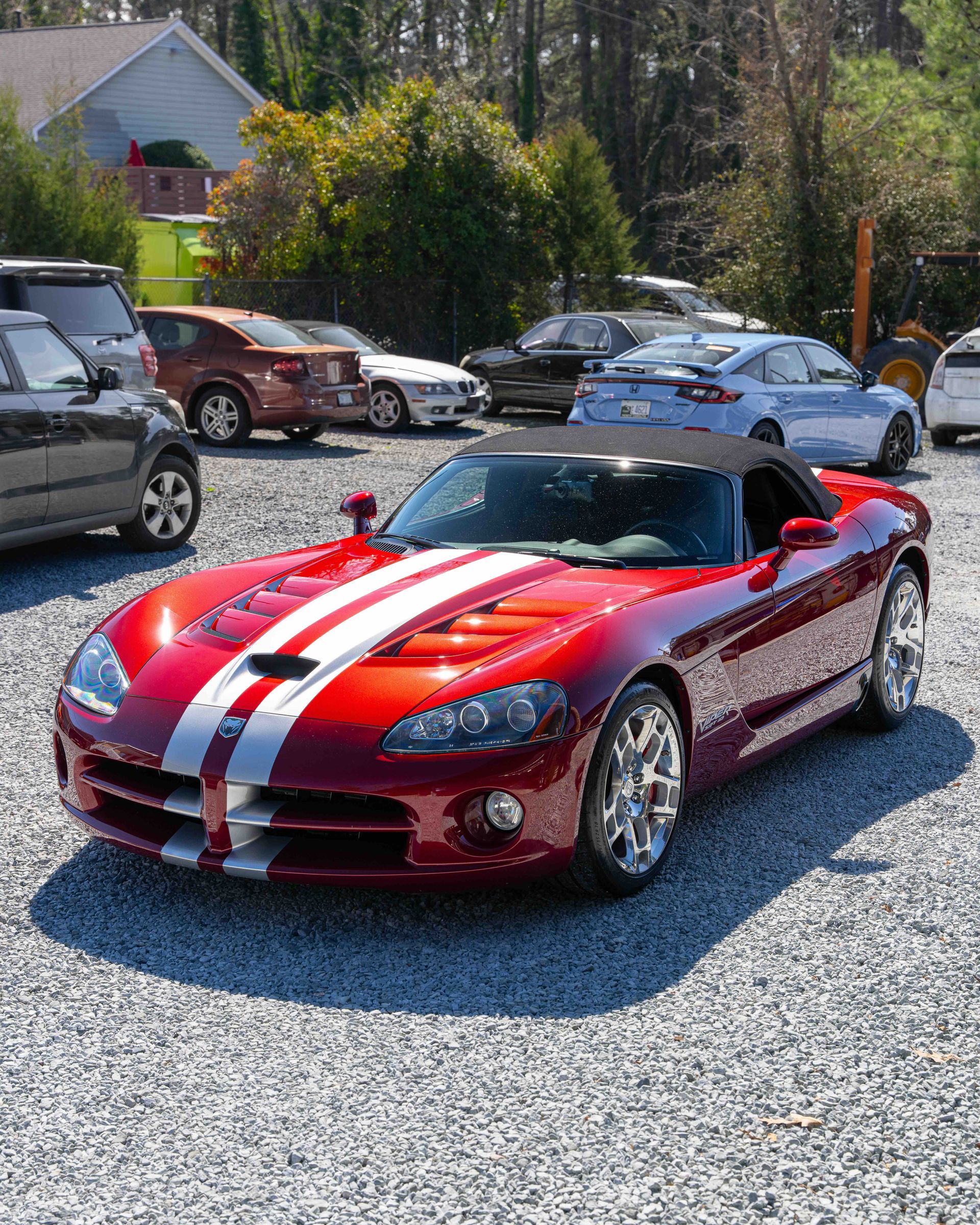 Red sports car with white racing stripes parked on a gravel lot among other vehicles.
