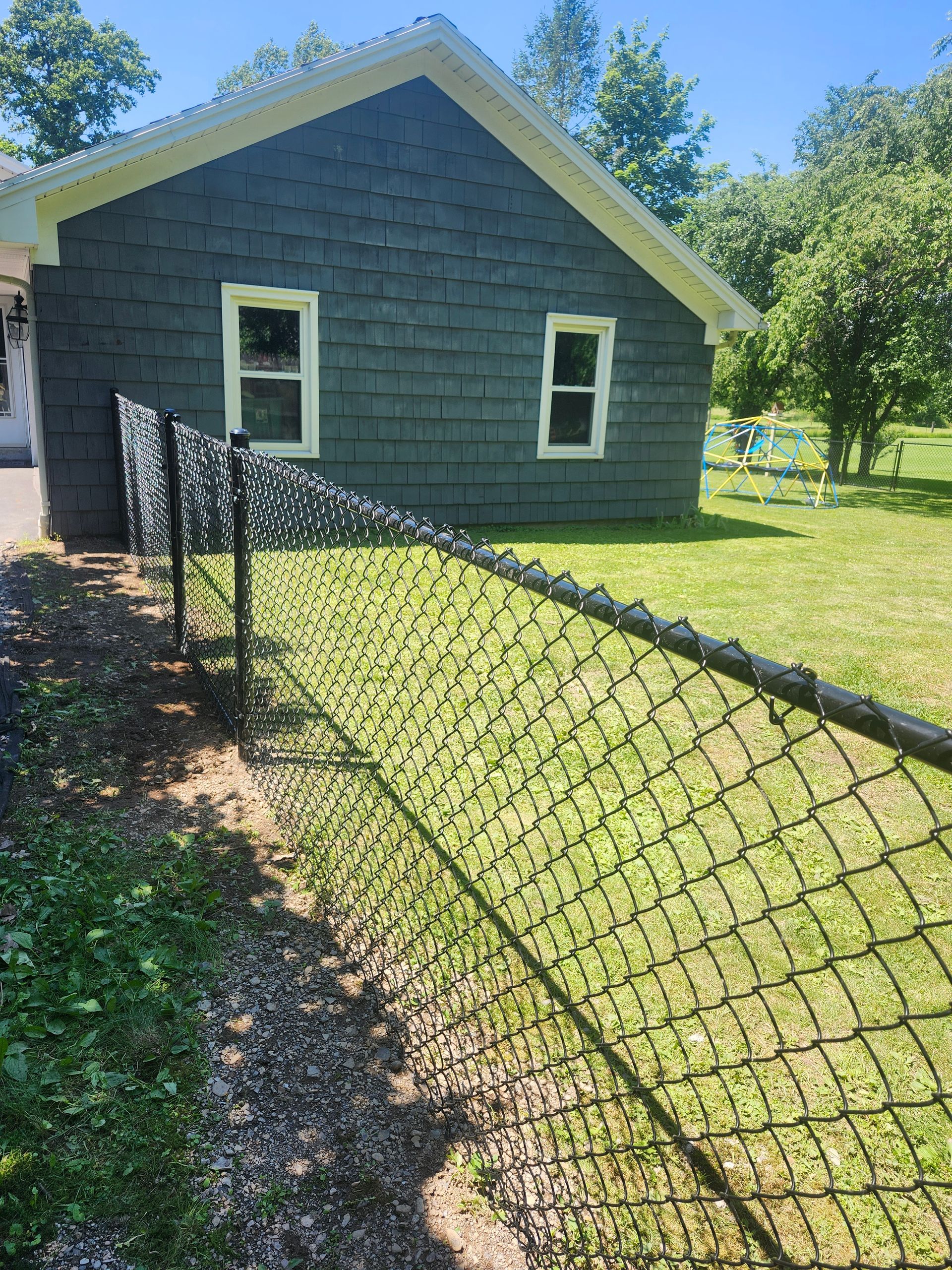 Chain-link fence leading to a gray house with two windows, set in a sunny grassy yard.