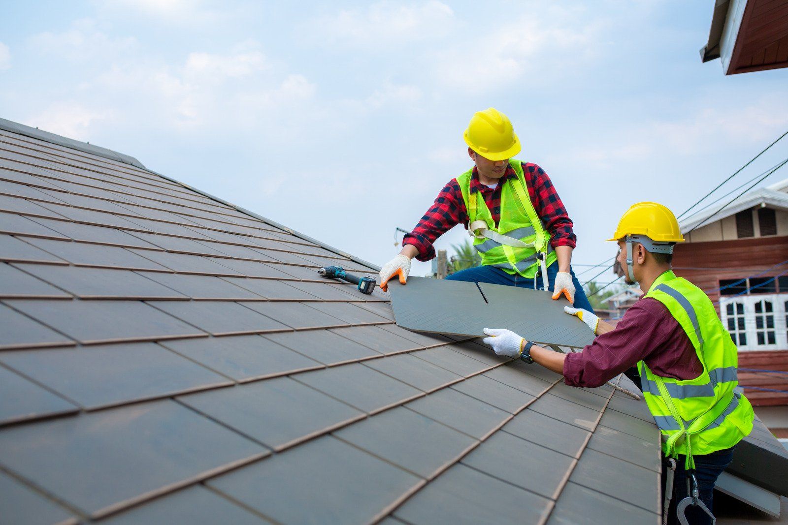 Two workers in hard hats and high-visibility vests install grey tiles on a sloped residential roof.