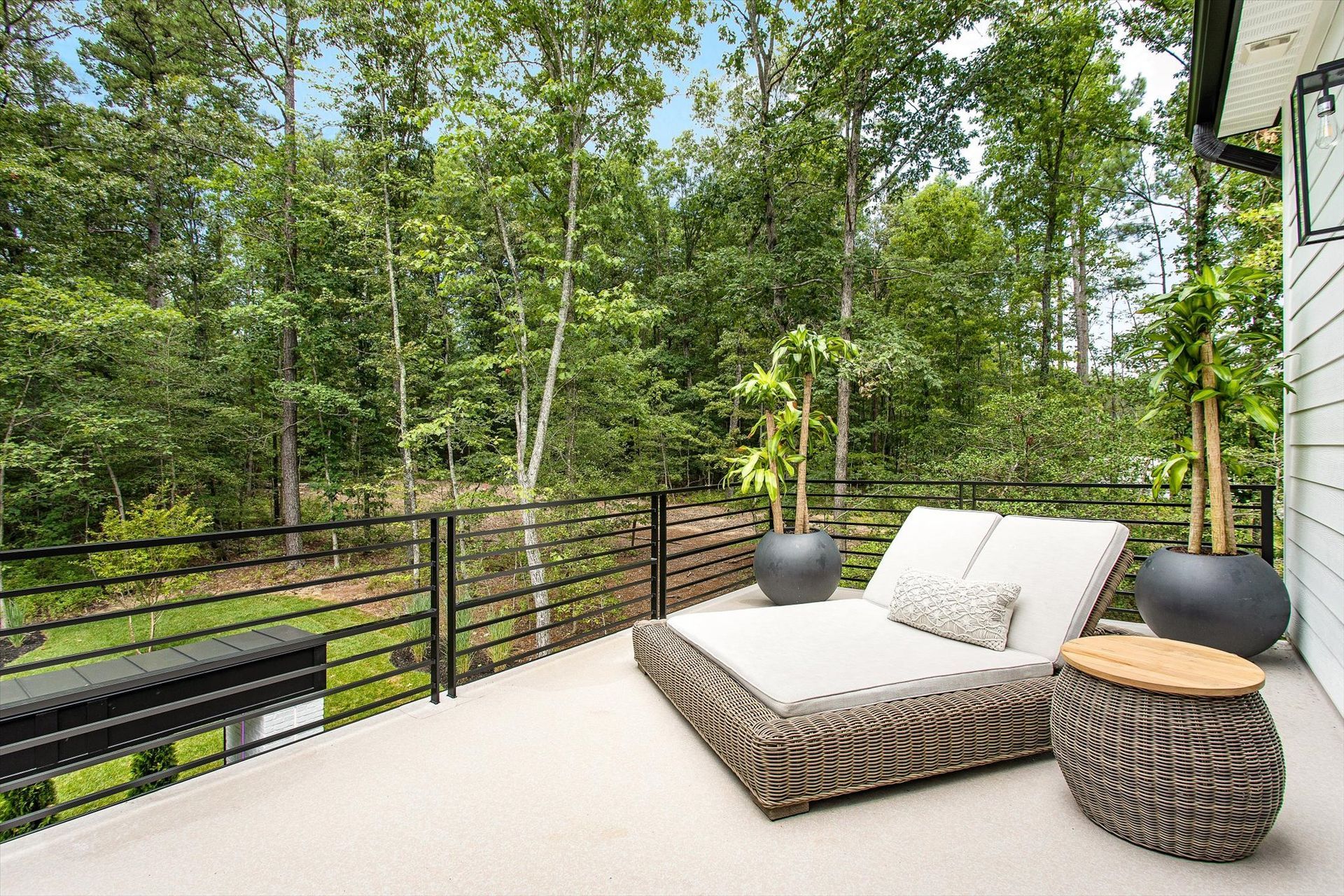 A patio with a bed , chairs , tables and potted plants surrounded by trees.