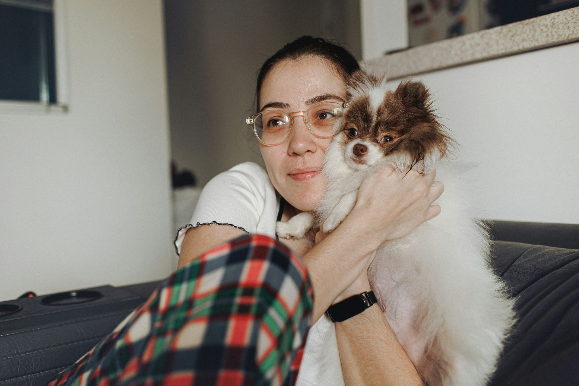 Woman holding a fluffy white-and-brown dog on a couch, wearing glasses and plaid pants