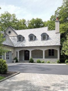 Large gray stone house with arched porch, dormer windows, and a paved driveway surrounded by trees