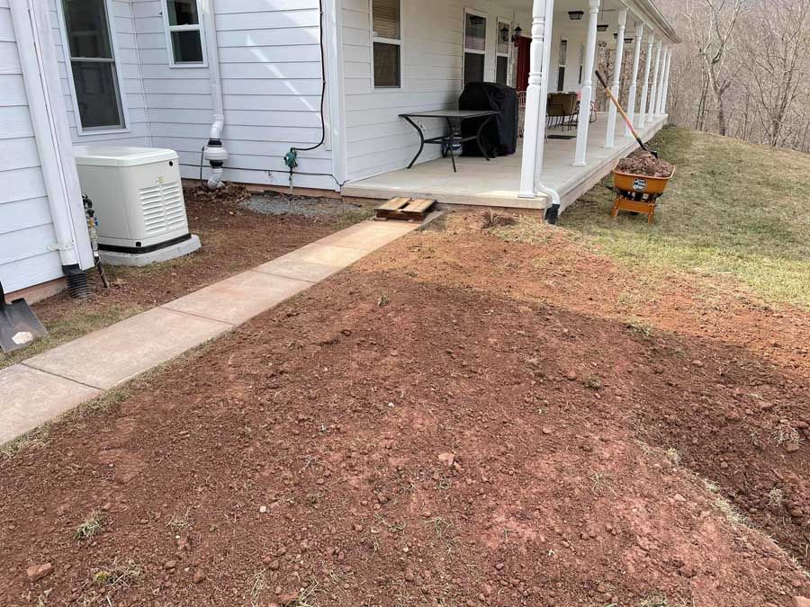 Front porch and walkway beside a house, with a bare dirt yard and a wheelbarrow on the right.