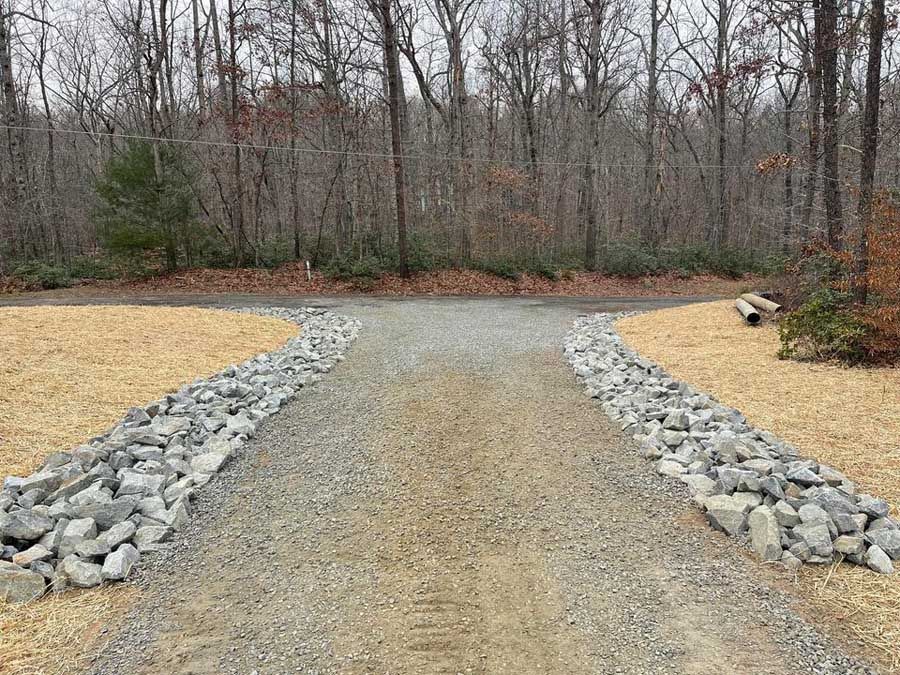 Gravel driveway bordered by rocks leading into a wooded area in late fall