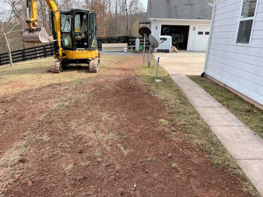 Mini excavator on a dirt yard beside a house and driveway, preparing a cleared path