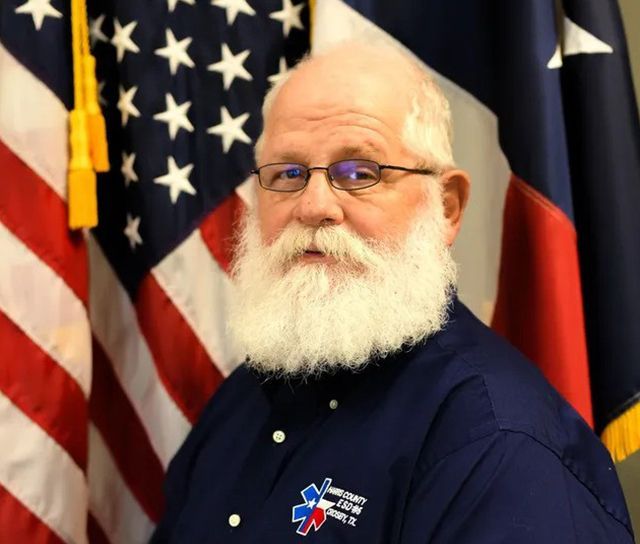 A man with a beard and glasses is standing in front of an American flag
