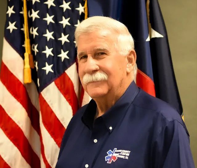 A man with a mustache is standing in front of an American flag.
