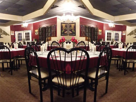 Restaurant interior with round tables set for dining, red tablecloths, black chairs, and dark red walls.