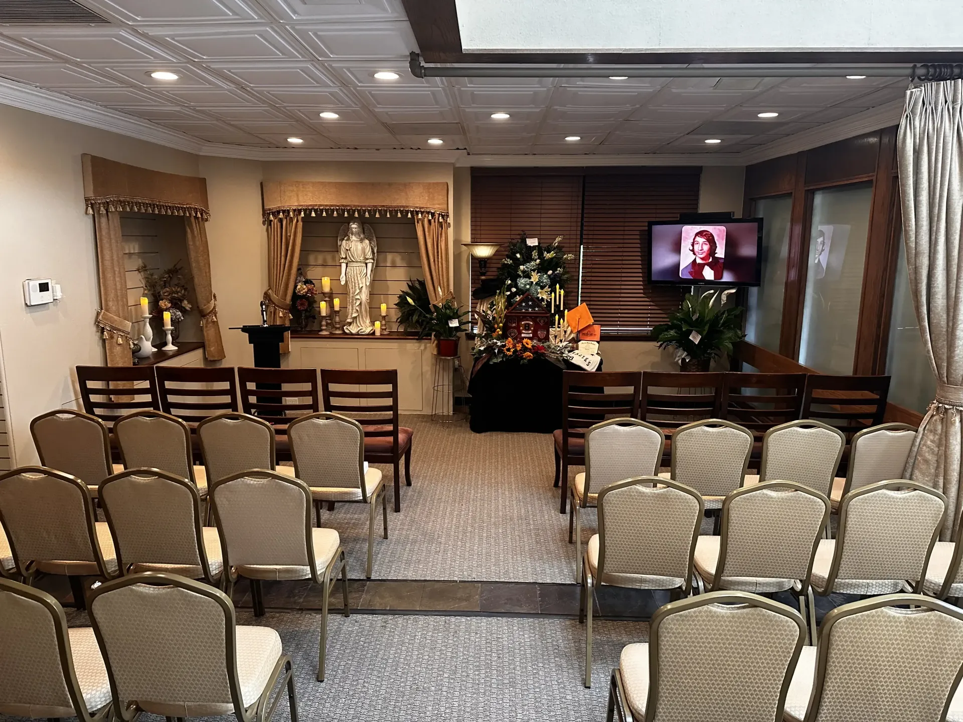 Indoor memorial service setup with rows of chairs facing a small altar and a screen showing a speaker.