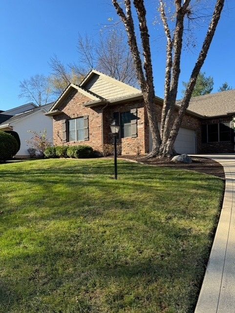 Brick house with tan roof, large tree, green lawn, and walkway on a sunny day.